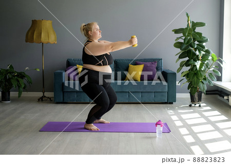 Young woman trains with yellow dumbbells and does squats at the same time. Concept of doing sports at home. Overweight lady training at home in front of sofa in room . Plus size strengthens biceps Young woman trains with yellow dumbbells and does squats at the same time. Concept of doing sports at home. Overweight lady training at home in front of sofa in room . Plus size strengthens biceps 88823823