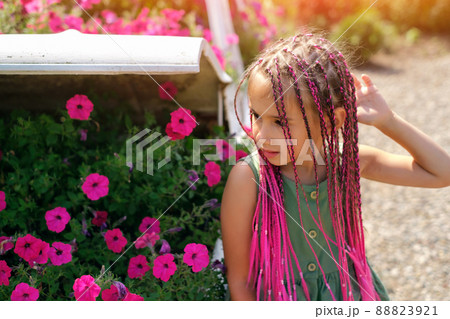 A beautiful cute girl with pink Afro-pigtails from kanekalon stands in front of a flower bed with flowers. a warm summer day. Petunia flowers with a pronounced sweet smell. zizi pigtails. 88823921
