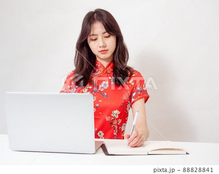Smiling young asian woman in traditional cheongsam qipao dress taking notes on notebook while using laptop isolated on white background. Smiling young asian woman in traditional cheongsam qipao dress taking notes on notebook while using laptop isolated on white background. 88828841