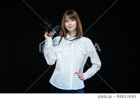 Portrait of a young woman wearing a white shirt holding a camera in her hand. The concept of a successful photographer. Isolated black background. 88833683