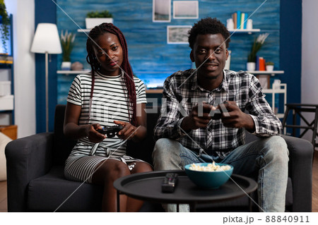 Relaxed african american couple playing video games on console while having a good time sitting at home on the sofa. Joyful people enjoying free time while having fun together in living room. Relaxed african american couple playing video games on console while having a good time sitting at home on the sofa. Joyful people enjoying free time while having fun together in living room. 88840911
