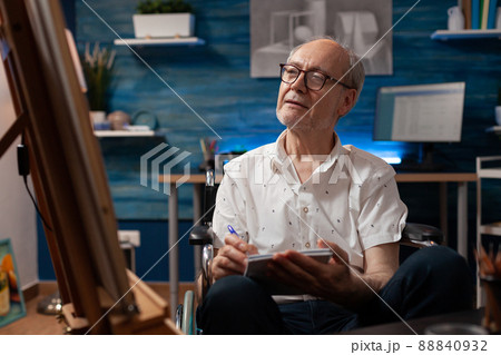 Elderly man artist living with disability doing sketch on notebook for practice before drawing on easel in home art studio. Creative senior hobby art creator taking notes keeping journal. Elderly man artist living with disability doing sketch on notebook for practice before drawing on easel in home art studio. Creative senior hobby art creator taking notes keeping journal. 88840932