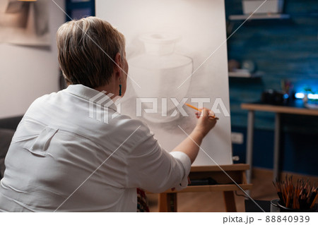 Over shoulder view of older woman drawing orginal masterpiece using sharp pencil on paper sketching vase model in home art studio. Portrait of elderly artist sketching still life in workshop. Over shoulder view of older woman drawing orginal masterpiece using sharp pencil on paper sketching vase model in home art studio. Portrait of elderly artist sketching still life in workshop. 88840939