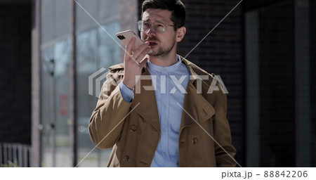 Stylish man making voice message or using virtual assistant app on mobile phone. Businessman in glasses recording voice message on speakerphone while walking near modern office building. Stylish man making voice message or using virtual assistant app on mobile phone. Businessman in glasses recording voice message on speakerphone while walking near modern office building. 88842206