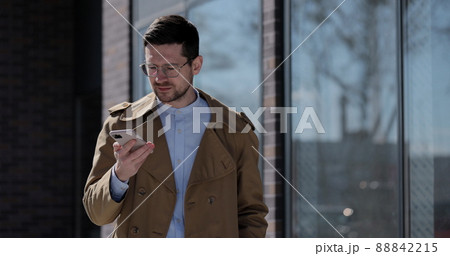 Stylish man making voice message or using virtual assistant app on mobile phone. Businessman in glasses recording voice message on speakerphone while walking near modern office building. Stylish man making voice message or using virtual assistant app on mobile phone. Businessman in glasses recording voice message on speakerphone while walking near modern office building. 88842215