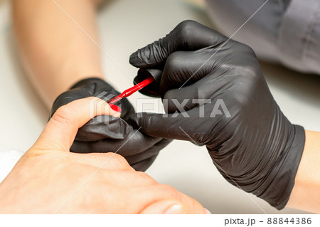 Manicure varnish painting. Close-up of a manicure master wearing rubber black gloves applying red varnish on a female fingernail in the beauty salon Manicure varnish painting. Close-up of a manicure master wearing rubber black gloves applying red varnish on a female fingernail in the beauty salon 88844386