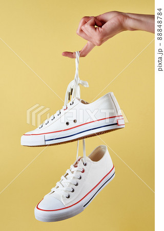 White female sport sneakers hanging on woman hand with shoe lace against yellow background, Casual trendy shoes closeup, Creative minimal design 88848784