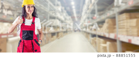 A warehouse worker points her finger up. Smiling young woman in work clothes in front of a huge warehouse 88851555