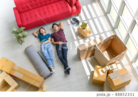 Top view of young asian couple smiling, relaxing and resting after unpacking cardboard boxes in new house with cardboard boxes on floor Top view of young asian couple smiling, relaxing and resting after unpacking cardboard boxes in new house with cardboard boxes on floor 88852331