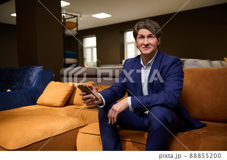Portrait of a mature European man shopping or working in upholstered furniture warehouse, sitting on a sofa with smartphone in his hands and looking at camera Portrait of a mature European man shopping or working in upholstered furniture warehouse, sitting on a sofa with smartphone in his hands and looking at camera 88855200