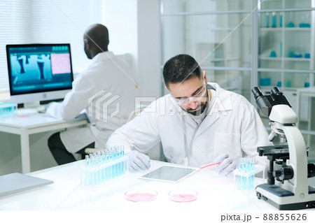 Young serious male chemist studying substance in petri dish by workplace 88855626