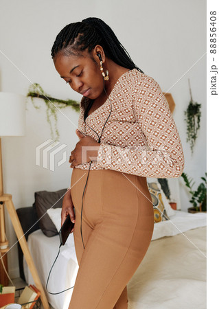 African pregnant woman using her mobile phone for enjoying music while standing in bedroom African pregnant woman using her mobile phone for enjoying music while standing in bedroom 88856408