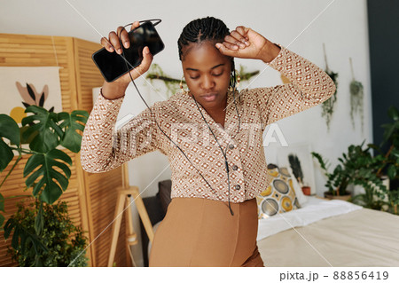 African pregnant woman in headphones dancing in bedroom while listening to music on her mobile phone 88856419