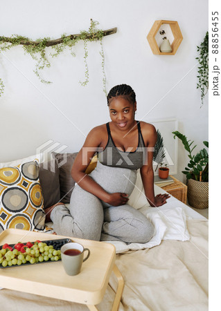 Portrait of African pregnant girl smiling at camera sitting on bed with fruits and tea on tray 88856455