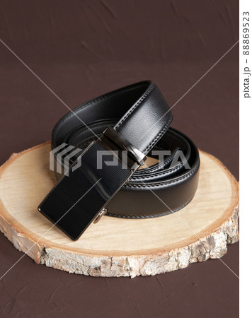 Rolled-up black fashionable men's leather belt with automatic buckle on wood slice on brown background. Male accessory. Front view, vertical photo. Rolled-up black fashionable men's leather belt with automatic buckle on wood slice on brown background. Male accessory. Front view, vertical photo. 88869523