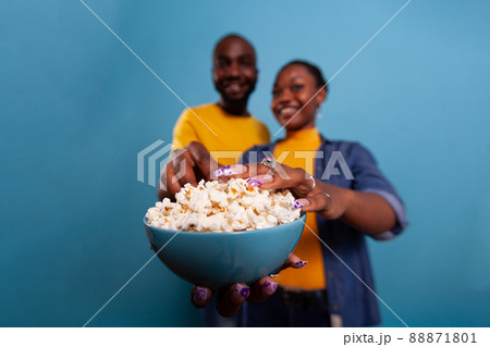 Joyful couple eating popcorn from glass bowl in studio, watching funny movie on television and having snack. Boyfriend and girlfriend enjoying food and comedy film on tv for entertainment. 88871801