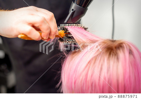 Drying short pink hair of young caucasian woman with a black hairdryer and black round brush by hands of a male hairdresser in a hair salon, close up Drying short pink hair of young caucasian woman with a black hairdryer and black round brush by hands of a male hairdresser in a hair salon, close up 88875971