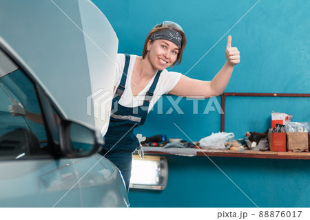 Portrait of a young smiling female mechanic in glasses and coveralls shows thumb up from behind the hood of a car. The concept of women's work in male professions Portrait of a young smiling female mechanic in glasses and coveralls shows thumb up from behind the hood of a car. The concept of women's work in male professions 88876017