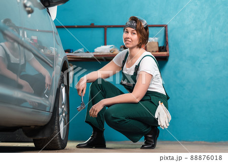 Young smiling female mechanic in blue coveralls squatting near tire and holding ratchet wrench. The concept of women's work in men's professions and equality Young smiling female mechanic in blue coveralls squatting near tire and holding ratchet wrench. The concept of women's work in men's professions and equality 88876018