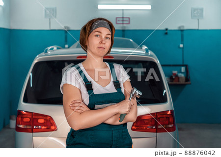 Portrait of a young caucasian plus-sized woman in overall holding a wrench. At the background a car in an auto repair shop. The concept of women's equality Portrait of a young caucasian plus-sized woman in overall holding a wrench. At the background a car in an auto repair shop. The concept of women's equality 88876042