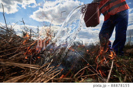 Firefighter ecologist fighting wildfire in field with cloudy sky on background. Close up of male environmentalist pouring water on burning dry grass. Natural disaster and ecology demage concept. Firefighter ecologist fighting wildfire in field with cloudy sky on background. Close up of male environmentalist pouring water on burning dry grass. Natural disaster and ecology demage concept. 88878155
