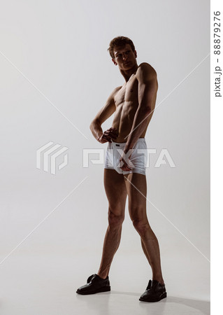 Portrait of young handsome shirtless sportive man wearing white boxer-briefs moves smoothly isolated on gray background. Wellness, fitness, beauty concept. Portrait of young handsome shirtless sportive man wearing white boxer-briefs moves smoothly isolated on gray background. Wellness, fitness, beauty concept. 88879276