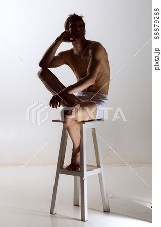 Looks sad, serious. One young shirtless sportive man in white boxer-briefs sitting on high chair isolated on grey background. Art, fashion, diversity, emotions 88879288