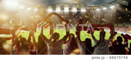 Victory. Back view of football, soccer fans cheering their team with scarfs at crowded stadium at evening time. Concept of sport, competition. Out of focus effect 88879748