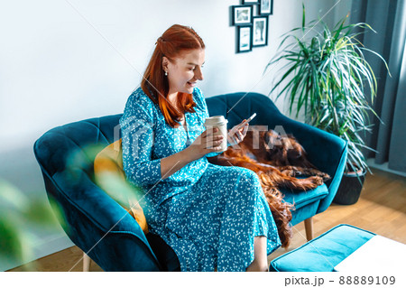 European happy 30s smile woman drinking coffee and used smartphone on a sofa at home. Big red irish setter dog sweet sleeping European happy 30s smile woman drinking coffee and used smartphone on a sofa at home. Big red irish setter dog sweet sleeping 88889109