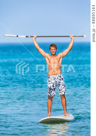 Happy watersport man having fun on stand-up paddleboard sup holding paddle up in the air in success on paddle board race. Handsome fit young athlete winning competition standing on surfboard Happy watersport man having fun on stand-up paddleboard sup holding paddle up in the air in success on paddle board race. Handsome fit young athlete winning competition standing on surfboard 88895163