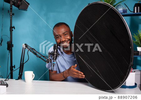 Portrait of photography equipment reviewer presenting beauty dish honeycomb grid in vlogging studio. Vlogger explaining features of studio flash light modifier sitting at desk with microphone. Portrait of photography equipment reviewer presenting beauty dish honeycomb grid in vlogging studio. Vlogger explaining features of studio flash light modifier sitting at desk with microphone. 88907206