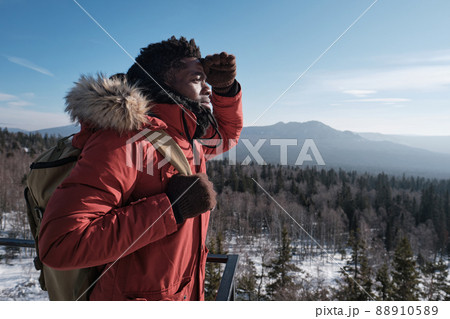 Young African American man wearing backpack standing at viewing platform on mountain top on winter day looking into distance 88910589