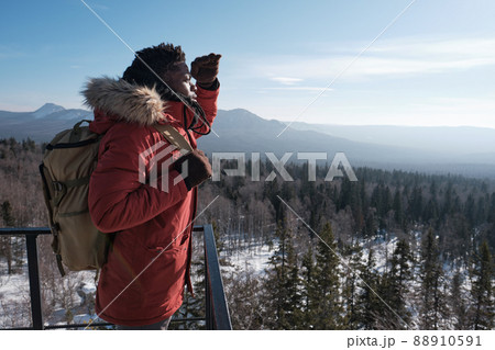Young African American traveler wearing backpack standing at observation deck on mountain top on winter day looking into distance 88910591