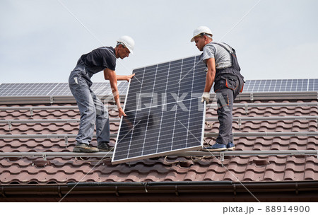 Men mounters lifting up photovoltaic solar modul on roof of house. Electricians in helmets installing solar panel system outdoors. Concept of alternative and renewable energy. 88914900