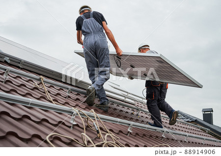 Men technicians carrying photovoltaic solar moduls on roof of house. Builders in helmets installing solar panel system outdoors. Concept of alternative and renewable energy. 88914906