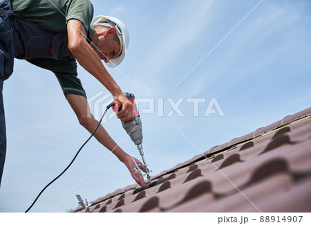 Worker prepearing to installation photovoltaic solar panel system on the roof of house. Man installer working with electric screwdriver, wearing uniform and helmet, on blue sky background. 88914907