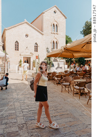 Girl Tourist Walking Through Ancient Narrow Street On A Beautiful Summer Day In MEDITERRANEAN MEDIEVAL CITY, OLD TOWN BUDVA, MONTENEGRO. Young Beautiful Cheerful Woman Walking On Old Street At Girl Tourist Walking Through Ancient Narrow Street On A Beautiful Summer Day In MEDITERRANEAN MEDIEVAL CITY, OLD TOWN BUDVA, MONTENEGRO. Young Beautiful Cheerful Woman Walking On Old Street At 88917547