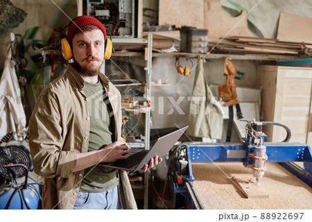 Horizontal medium portrait of young Caucasian man working in carpentry workshop holding laptop looking at camera Horizontal medium portrait of young Caucasian man working in carpentry workshop holding laptop looking at camera 88922697