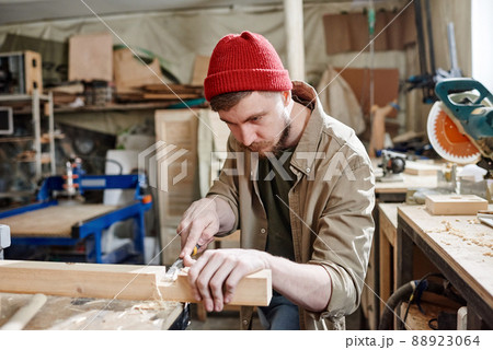 Bearded Caucasian carpenter wearing red knit cap cutting mortise in wood plank using chisel 88923064