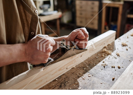 Medium close-up of unrecognizable carpenter working in workshop using hand plane to shape and smooth wood plank surface Medium close-up of unrecognizable carpenter working in workshop using hand plane to shape and smooth wood plank surface 88923099