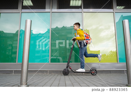 Modern teenager with backpack rides on electric scooter on brick wall background. Boy comes back from school 88923365