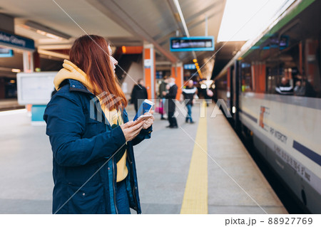 Young redhhead woman waiting on station platform with backpack on background electric train using smart phone. Railroad transport concept, Traveler 88927769