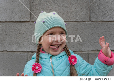 A portrait of a cheerful child against a gray stone wall. A five-year-old girl with her tongue out. A gesticulating daughter with long pigtails. 88943941