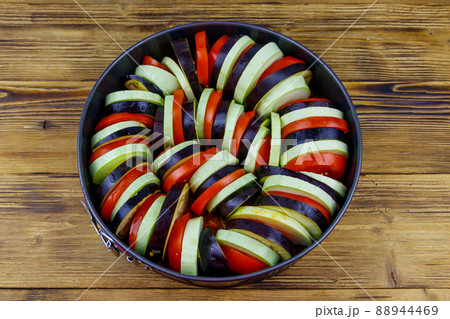 Preparing of ratatouille. Raw vegetables for ratatouille in round baking dish on wooden table. Traditional French vegetable dish Preparing of ratatouille. Raw vegetables for ratatouille in round baking dish on wooden table. Traditional French vegetable dish 88944469