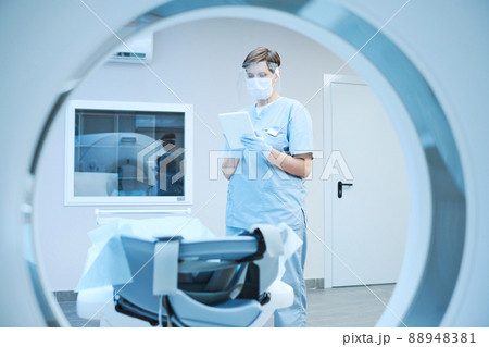 Serious nurse in protective mask and gloves using tablet and reading patients card while preparing MRI scanner for covid patient Serious nurse in protective mask and gloves using tablet and reading patients card while preparing MRI scanner for covid patient 88948381