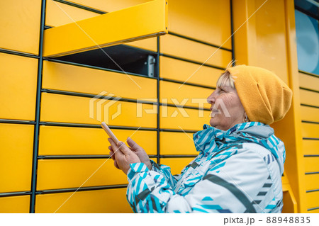 Young caucasian woman getting parcel from cell of automatic post terminal outdoors.  88948835