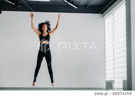 A happy woman in black sportswear makes a jump up on a white background. Girl jumping raises her hands up in the gym 88954268