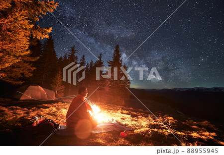 Side view of alone hiker, warming himself by campfire on mat, looking at surrounding mountain landscapes. Luxurious starry sky, Milky Way galaxy over mountain hills. Side view of alone hiker, warming himself by campfire on mat, looking at surrounding mountain landscapes. Luxurious starry sky, Milky Way galaxy over mountain hills. 88955945