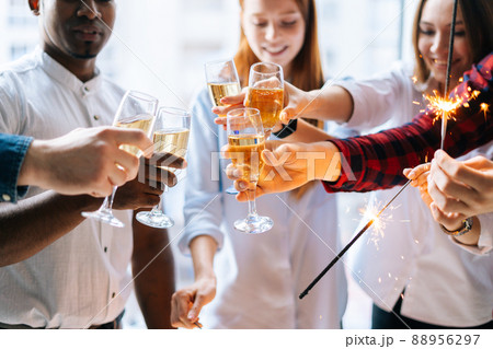 Close-up cropped shot happy business team of young multiracial businesspeople holding Bengal lights and clinking glasses of champagne at corporate party, on background of window and cityscape. Close-up cropped shot happy business team of young multiracial businesspeople holding Bengal lights and clinking glasses of champagne at corporate party, on background of window and cityscape. 88956297