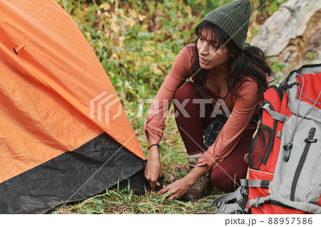 Young mixed race woman in hat setting up orange tent in forest while preparing for camping 88957586